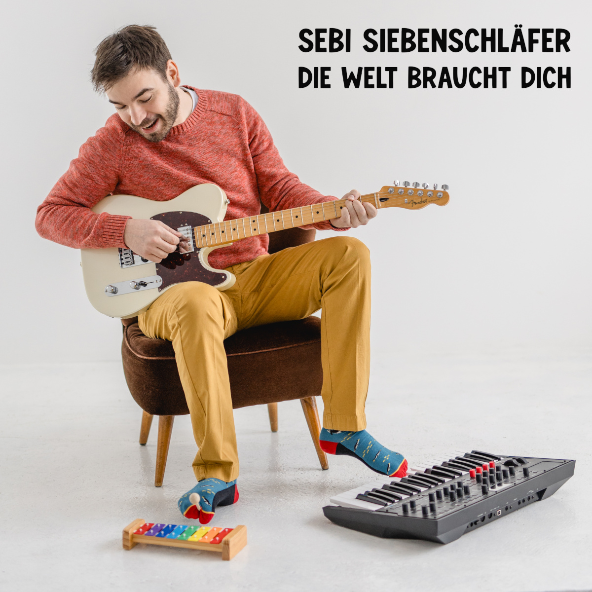 Cover des Albums DIE WELT BRAUCHT DICH von Sebi Siebenschläfer. Darauf zu sehen ist Sebi Siebenschläfer, der vor weißem Hintergrund auf einem Sessel sitzt, mit den Händen elektrische Gitarre spielt, mit dem einen Fuß ein Keyboard, und mit dem anderen Fuß ein Glockenspiel.